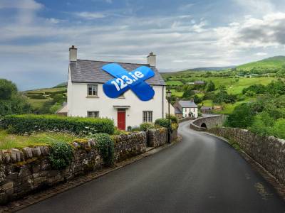 A traditional white Irish cottage with a red door on a winding country road, featuring a blue 123.ie "cover that sticks" bandage logo on the roof.